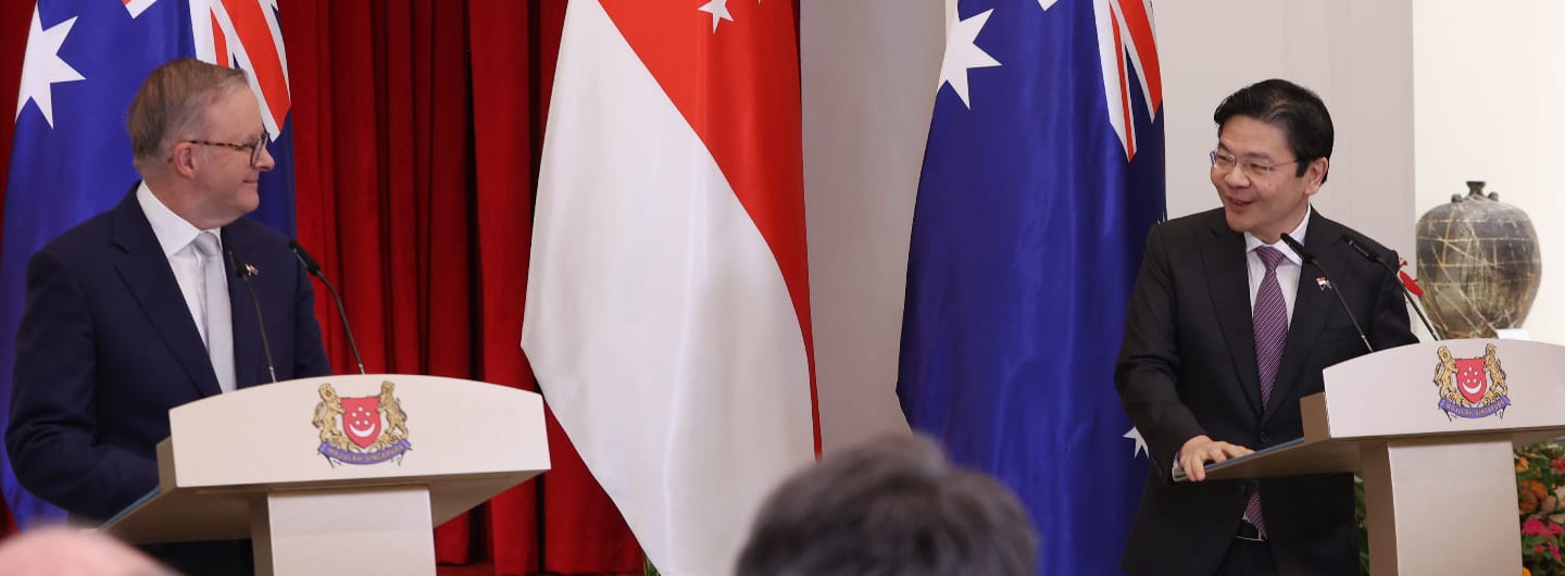 Two men at podiums flanking Singapore & Australia flags, smiling. Podium logos are present.